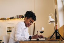 Side view low angle of African American female writing in notebook while watching online tutorial via mobile phone at home — Stock Photo