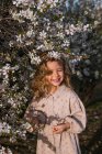 Liebenswert lächelndes kleines Kind im Kleid steht neben blühendem Baum mit Blumen im Frühlingspark und schaut weg — Stockfoto