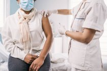 Обрезанная неузнаваемая женщина-врач в защитной форме, латексных перчатках и маске для лица, прививающая анонимную афро-американскую зрелую пациентку в клинике во время вспышки коронавируса — стоковое фото