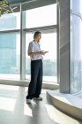 Jovem solitária e pouco emocional de pé no escritório vazio com grande janela navegando no telefone celular — Fotografia de Stock
