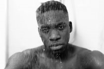 Noir et blanc de jeune homme noir sans émotion prenant une douche dans la salle de bain légère et regardant la caméra et l'eau sur le visage — Photo de stock