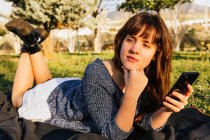 Vista laterale della donna spensierata sdraiata sul plaid sul prato e utilizzando lo smartphone mentre si gode il picnic nella giornata di sole in primavera — Foto stock