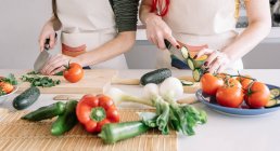 Cultivo irreconocible homosexual novias cortar pepino mientras se prepara comida vegetariana saludable en la mesa en casa - foto de stock
