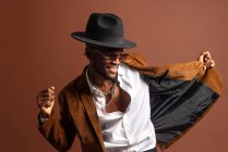 Joyeux jeune homme afro-américain dans des vêtements à la mode et chapeau dansant en regardant loin avec sourire dents — Photo de stock