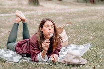 Mujer despreocupada tumbada sobre una manta en el bosque y soplando diente de león mientras disfruta de un picnic en Australia - foto de stock