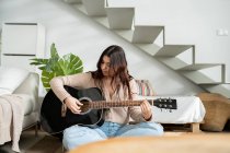 Молодая женщина-музыкант играет на акустической гитаре, сидя со скрещенными ногами на полу дома — стоковое фото
