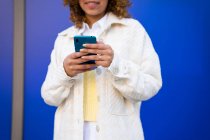 Ritagliato irriconoscibile felice femmina afroamericana utilizzando smartphone mentre in piedi su sfondo blu — Foto stock