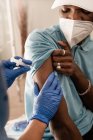 Médico de corte em uniforme protetor e luvas de látex vacinando paciente afro-americano masculino irreconhecível na clínica durante o surto de coronavírus — Fotografia de Stock