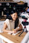 Jovem étnica alegre feminino vlogger com notebook sentado à mesa com câmera de fotos no tripé na cozinha — Fotografia de Stock