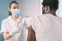 Especialista médica em uniforme de proteção, luvas de látex e máscara facial vacinando pacientes afro-americanos na clínica durante o surto de coronavírus — Fotografia de Stock