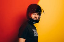 Вид збоку на впевненого брутального дорослого чоловіка в шоломі на чорному мотоциклі, який дивиться на камеру, стоячи на тлі барвистого червоного і жовтого фону — стокове фото