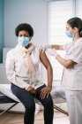 Female medical specialist in protective uniform, latex gloves and face mask vaccinating African American mature woman patient in clinic during coronavirus outbreak — Stock Photo