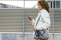 Vista lateral de la optimista mujer afroamericana con peinado afro navegando en el teléfono inteligente mientras está de pie contra la pared metálica en el área urbana de la ciudad - foto de stock