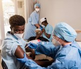 Médica em uniforme de proteção e luvas de látex vacinando paciente afro-americano na clínica durante o surto de coronavírus — Fotografia de Stock