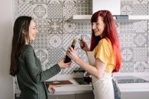 Content young homosexual girlfriends with bottle of wine speaking while looking at each other between stove and fresh vegetables — Stock Photo