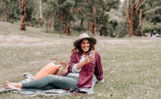 Веселая женщина в шляпе сидит на одеяле на лугу в лесу и просматривает мобильный телефон, наслаждаясь пикником в Австралии — стоковое фото