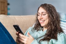 Молода щаслива усміхнена жінка з бірюзовим светром і окулярами, лежачи на дивані за допомогою мобільного телефону — стокове фото