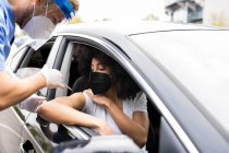 Side view of male doctor in protective uniform, latex gloves and face shield vaccinating African American female patient inside the car on a drive through mobile clinic during coronavirus outbreak — Stock Photo