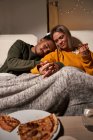 Pareja multiétnica sentada en sofá y novia comiendo deliciosa pizza mientras su novio duerme - foto de stock