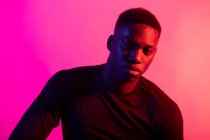 Joven hombre negro confiado en traje deportivo oscuro mirando a la cámara sobre fondo rosa neón en estudio oscuro - foto de stock