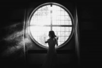 Back view of unrecognizable gentle female looking over the shoulder touching fence on round shaped window in house in sunlight — Stock Photo