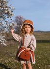 Menina adorável em vestido e chapéu de pé perto da árvore com flores florescentes e olhando para a câmera no jardim da primavera — Fotografia de Stock
