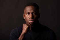 Confiante jovem afro-americano cara em roupas pretas com a mão no queixo olhando para a câmera no fundo preto no estúdio de luz — Fotografia de Stock
