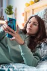 Молода жінка з бірюзовим светром і окулярами лежить на ліжку за допомогою мобільного телефону — стокове фото