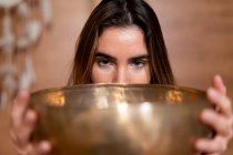 Crop young female covering mouth with Himalayan bowl while looking at camera on blurred background — Stock Photo