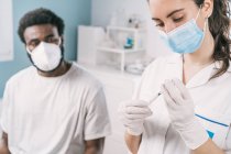 Female doctor in latex gloves and face shield filling in syringe from bottle with vaccine preparing to vaccinate unrecognizable African American man patient in clinic during coronavirus outbreak — Stock Photo