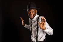 Young masculine ethnic male model in hat and trousers standing dancing while looking at camera on black background — Stock Photo