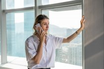 Joven mujer de negocios de pie en la oficina con grandes ventanas que tienen una llamada telefónica en el teléfono móvil - foto de stock