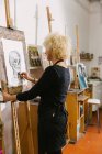 Side view of talented female artist standing at easel and drawing on paper with pencil in art studio — Stock Photo