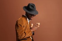 Seitenansicht eines jungen Afroamerikaners in trendiger Kleidung und Hut, der auf braunem Hintergrund wegschaut — Stockfoto
