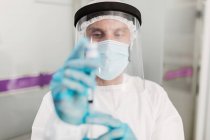 Розмиті невизначені ліки в захисній масці для обличчя та латексні рукавички з флаконом коронавірусної вакцини та шприцом, що показують камеру, стоячи в лікарняній кімнаті — стокове фото