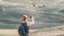 Vista posterior de una mochilera anónima tomando fotos de colinas en el teléfono celular durante el viaje de verano a la luz del sol - foto de stock
