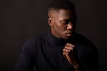 Уверенный молодой афроамериканец в черной одежде с закрытыми глазами на черном фоне в светлой студии — стоковое фото