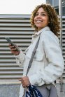 Mujer afroamericana optimista con peinado afro navegando en el teléfono inteligente mientras está de pie contra la pared metálica en el área urbana de la ciudad - foto de stock