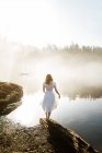 Mujer vista trasera vestida con un vestido blanco en una roca mirando a un lago en un día de niebla - foto de stock