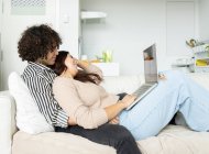 Frau mit Netbook spricht mit tätowiertem Freund, während sie auf der Couch im Hauszimmer auf den Bildschirm schaut — Stockfoto