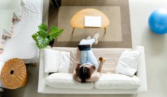 De cima jovem fêmea com tablet e xícara de bebida quente descansando com pernas cruzadas no sofá na sala de estar — Fotografia de Stock