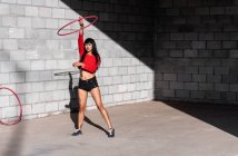 Jovem mulher tatuada em activewear girando hula hoop enquanto dança contra paredes de tijolo com sombras e olhando para a frente na luz do sol — Fotografia de Stock