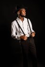 Giovane modello maschile etnico maschile maschile in cappello e pantaloni in piedi mentre distoglie lo sguardo su sfondo nero con il fumo — Foto stock