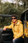 Vista lateral del aventurero con gafas de sol y chaqueta amarilla junto a su coche todoterreno - foto de stock
