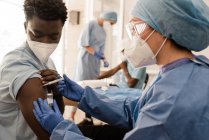 Médecin féminin en uniforme de protection et gants de latex vaccinant le patient afro-américain masculin en clinique pendant l'épidémie de coronavirus — Photo de stock