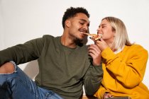 Positivo multirracial casal comer pizza fatia juntos enquanto se divertindo e olhando para o outro — Fotografia de Stock