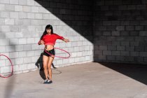 Joven mujer tatuada en ropa deportiva girando hula hoop mientras baila contra paredes de ladrillo con sombras - foto de stock