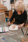 Alto angolo di artista femminile nella pittura grembiule con acquerelli su carta mentre seduto a tavola in laboratorio creativo — Foto stock