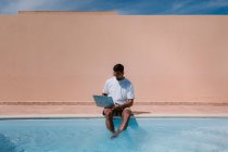 Чоловічий фрілансер в сонцезахисних окулярах сидить біля басейну і переглядає нетбук під час роботи віддалено на проекті під час літніх канікул — стокове фото