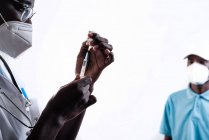 Этнический врач заполняет шприц из бутылки вакциной, готовящейся к вакцинации афроамериканского пациента на белом фоне в клинике во время вспышки коронавируса — стоковое фото